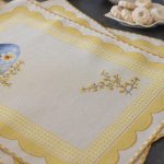 Tovaglietta pasquale in tessuto Gobelin con decorazioni floreali e un uovo blu ornato da fiori e rametti di mimosa. Il bordo smerlato e il motivo a quadretti gialli aggiungono un tocco raffinato ed elegante, perfetto per decorare la tavola di Pasqua con stile primaverile.