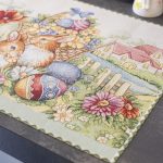 Tovaglietta pasquale in tessuto Gobelin con illustrazione di due coniglietti circondati da fiori primaverili, uova decorate e un cesto colmo di mimose. Lo sfondo raffigura una casetta di campagna con un ruscello e un giardino fiorito, creando un'atmosfera accogliente e festiva. Perfetta per decorare la tavola di Pasqua con eleganza e colori vivaci.